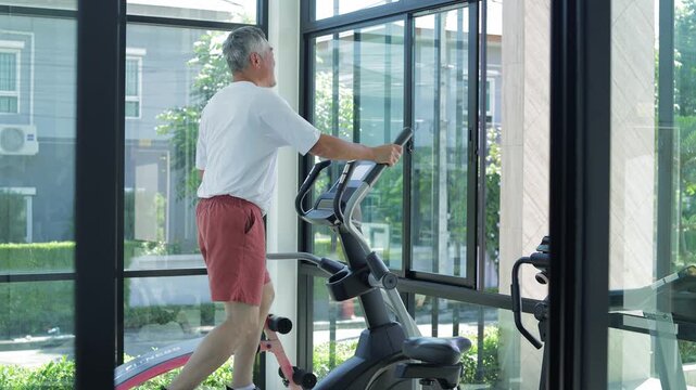 Senior Asian man exercising on an elliptical trainer inside a bright, modern glass-walled gym room, focusing on active retirement, healthy lifestyle, and cardiovascular fitness for seniors.