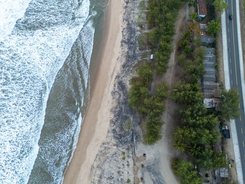 Aerial view of the coastline in Pacitan featuring sandy beach, crashing waves, lush trees, and a nearby road in East Java, Indonesia.