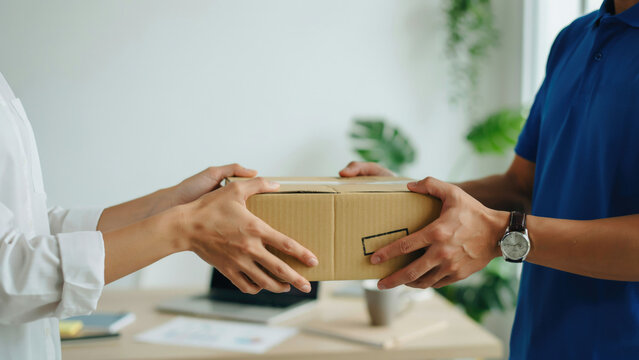 A courier in a blue shirt hands a cardboard parcel to a recipient in a white shirt inside an office environment with customer office setting delivery