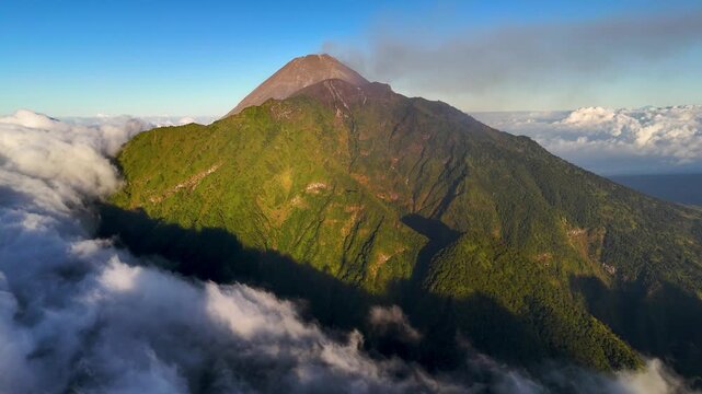 Aerial view of Mount Merapi with lush green slopes and volcanic smoke rising above the clouds in Selo, Central Java, Indonesia.