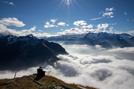 View of Stoos mountain ridge with a person sitting and looking over a sea of clouds towards snow-capped peaks under a bright sun in Morschach, Schwyz, Switzerland.