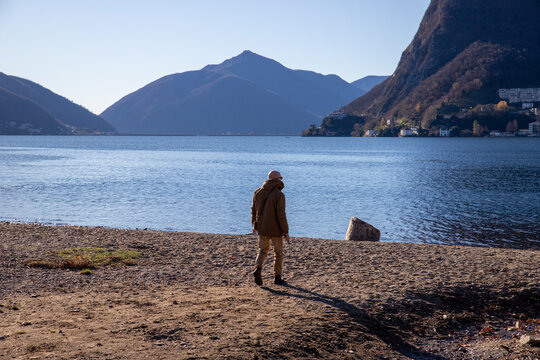 View of a man walking along the pebble beach of Lake Lugano with mountains in the background under a clear blue sky in Lugano, Ticino, Switzerland.