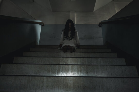 Halloween horror scene showing scary woman in white dress sitting on concrete stairs inside dark building, creating creepy atmosphere for spooky holiday celebration concept
