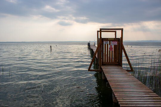View of a wooden pier extending into the calm waters of Lake Garda, featuring a gated entrance with warning signs under a cloudy sky in Lombardia, Italy.