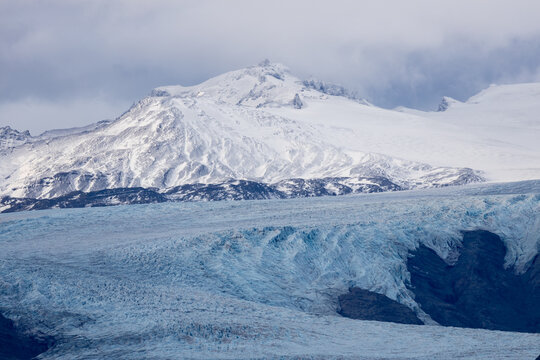 View of a massive glacier with deep blue ice textures and snow-covered mountain peaks under a cloudy sky in Iceland.