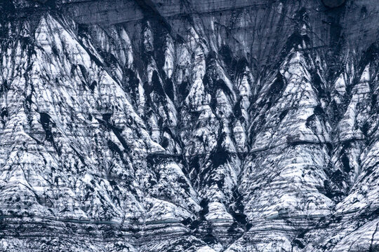 View of the intricate textures of a glacier surface featuring layers of white ice and dark volcanic ash deposits in Iceland.