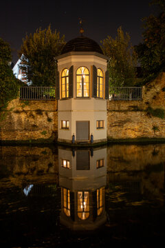 View of an illuminated historic pavilion reflecting in the calm canal water at night in Bruges, Flanders, Belgium.