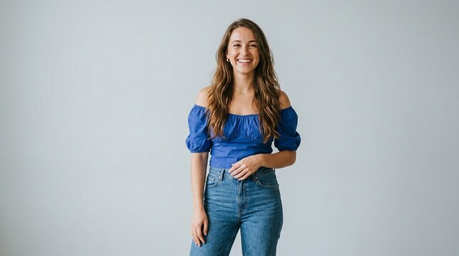 Smiling young woman with long brown hair wearing a blue off-shoulder blouse and jeans, standing confidently against a plain light gray studio background, cheerful portrait, casual fashion, bright