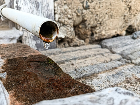 Close-up of an old white water pipe with rust and a single water droplet, set against weathered stone steps, highlighting aging infrastructure and water conservation.