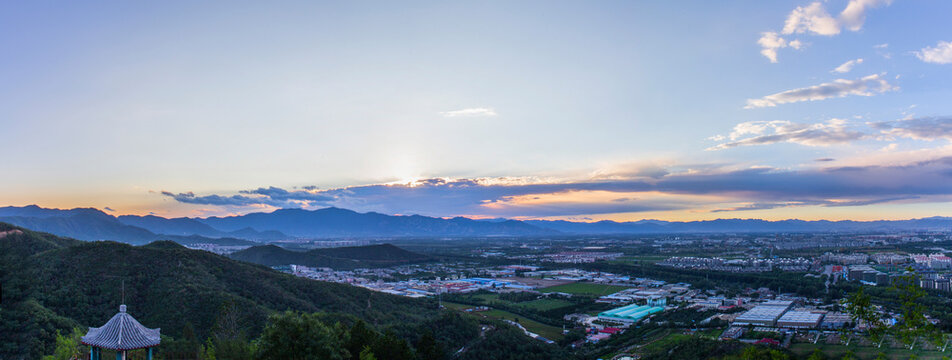 A panoramic view of a valley nestled between mountain ranges at dawn or dusk, with scattered settlements and greenery under a partly cloudy sky.