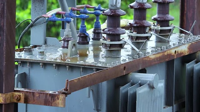 Power transformer substation shows rain soaked ceramic insulators and bushings on rusty metal frame, electrical infrastructure closeup.