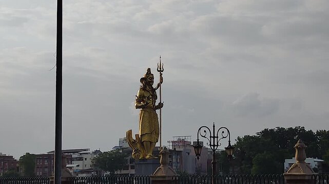 Sursagar Lake Vadodara features golden Shiva statue holding trident closeup with city buildings and lamp post below, Gujarat India.