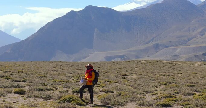 Annapurna Circuit, Mustang, Nepal, drone orbit shot circling a lone backpacker standing on a dry Himalayan desert plateau checking a map wearing a red hat and carrying a backpack