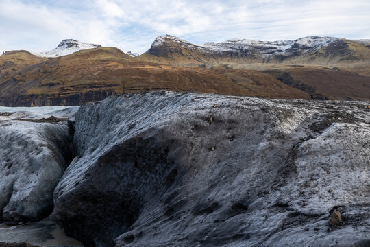 View of a glacier with dark, ash-covered ice and snow-capped mountains under an overcast sky in Iceland.