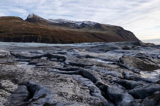 View of the rugged surface of a glacier with deep crevasses and dark volcanic ash deposits, set against a backdrop of snow-dusted mountains in Iceland.