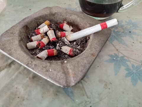 Close-up of a square ashtray filled with cigarette butts and ash, with a glass cup of coffee beside it on a patterned tabletop; candid scene of a smoking ritual and morning break.