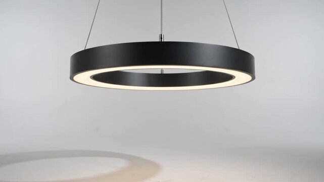 Pendant light fixture in a modern circular design hangs against a neutral studio background casting a soft glow below.
