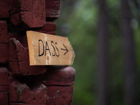 Handmade wooden sign with the Swedish word &ldquo;DASS&rdquo; and an arrow pointing toward an outdoor toilet, mounted on the corner of a traditional red timber building in a forest setting.