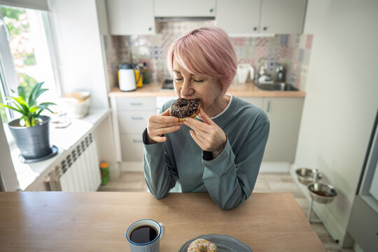 Mature woman bites chocolate donut during intense PMS craving, illustrating dietary relapse, sugar addiction and return to sweet pastries after unsuccessful effort to quit sugary comfort food.