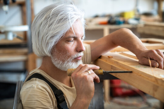 Concentrated professional craftsman restoring antique wooden furniture in carpentry workshop, thoughtful woodworking master at workstation using chisel instrument for precise carving of wood layers.