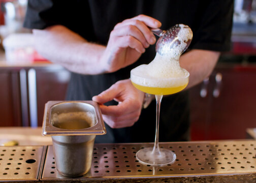 Serving cocktail. Mixologist work. Bartender adding frothy top ingredient from metal container to yellow liquid stemmed glass on counter.