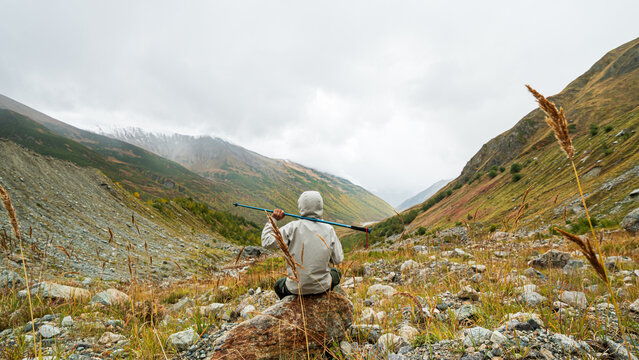 Hiker sitting on rock holding trekking pole over shoulders looking at autumn mountain valley near Shkhara glacier in Georgia.