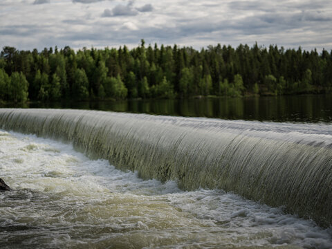 Water flowing over the low dam at Spegeldammen in Jokkmokk, Swedish Lapland, surrounded by green boreal forest under a cloudy summer sky.