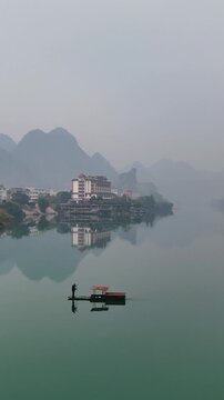 Vertical Video: Early morning mist over karst river with fisherman in wooden boat casting net on the water