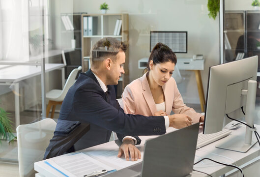 Colleagues in office conduct planning and analysis. Two managers review papers on a desktop computer monitor, pointing to details and updating tasks. Clear theme of teamwork and collaboration.
