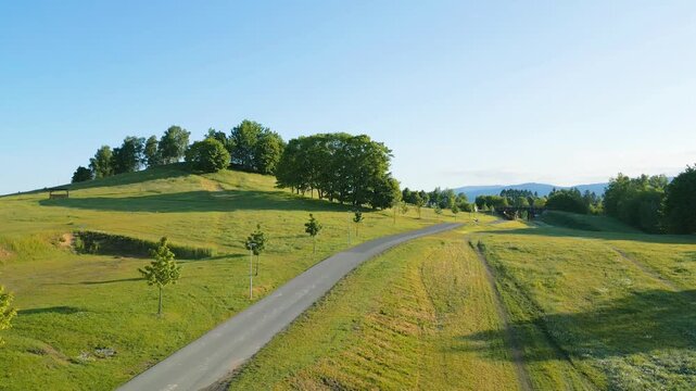 Aerial view of Cross-country ski area Vesec featuring green hills, a paved path, and trees under a clear blue sky in Liberec, Liberec Region, Czechia.