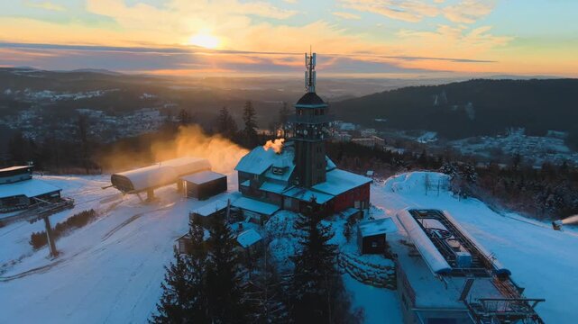 Aerial view of Tanvaldsky Spicak observation tower and ski resort with snow-covered slopes and ski lifts during sunset in Tanvald, Liberecky kraj, Czechia.