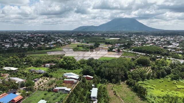 Slow forward drone push toward Ba Den Mountain across a rural town, flooded rice paddies and orchards opening in the foreground under bright cumulus cloud, Tay Ninh Province, Vietnam.