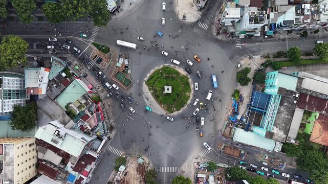 Top down aerial holding steady above a circular roundabout ringed by construction hoarding, motorbikes taxis and a bus flowing around the planted island, Ho Chi Minh City, Vietnam.