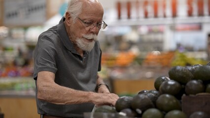 An elderly man examines avocados while shopping for groceries in a grocery store. He focuses on...