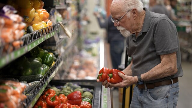 An elderly man is shopping for red bell peppers in a grocery store aisle surrounded by fresh vegetables and fruits