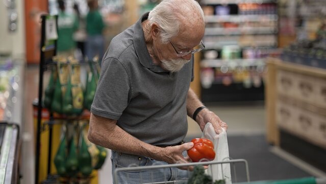 An elderly man examines a red bell pepper while shopping in a grocery store. The setting is busy with shoppers around