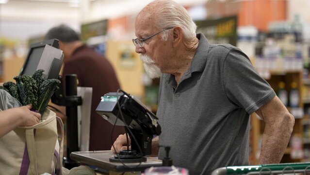 An elderly man stands at the checkout in a grocery store looking at items with focus. He interacts with the cashier and prepares to pay