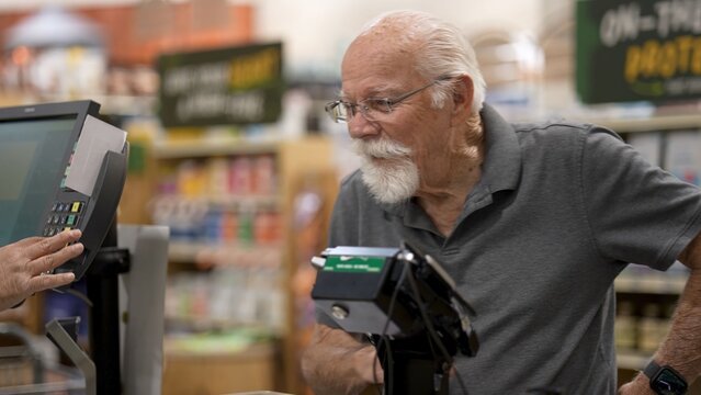 Elderly man stands at checkout in grocery store with items ready for purchase while interacting with the cashier
