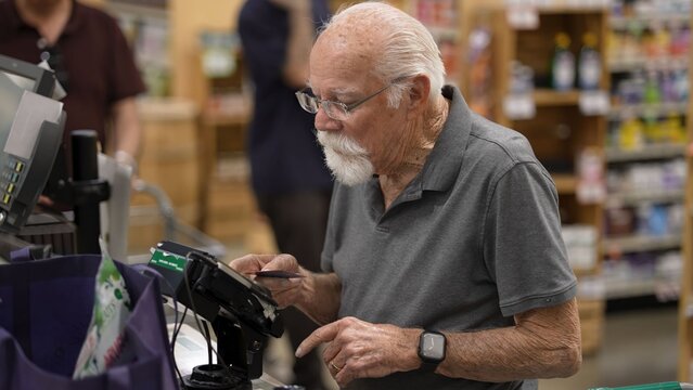 An elderly man shops in a grocery store. He stands at the checkout while using a card to pay for his items
