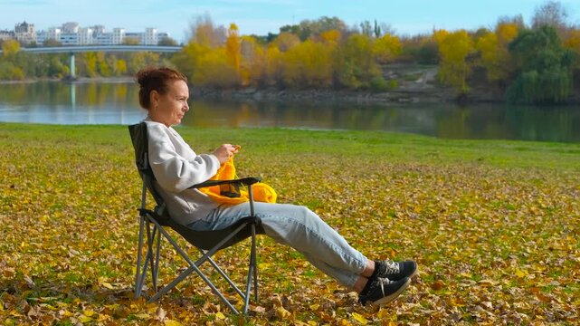 Woman knitting yarn relaxing in autumn park. Happy woman knitting with yellow yarn while sitting on a chair in a scenic autumn park by the river, enjoying a peaceful and creative outdoor hobby