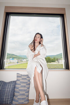 Woman wearing white bathrobe standing in the room with the window on large window.