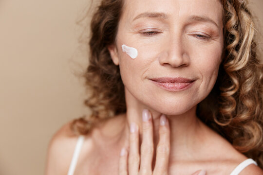 Smiling middle aged woman with natural skin applying white cream to her cheek. Close up on a beige background. Concept of skincare, wellness, selfcare, and authentic beauty.
