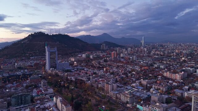 Aerial view of the sprawling Santiago city skyline with Cerro San Cristobal and distant mountains under a dramatic cloudy sky at dusk in Santiago, Santiago Metropolitan, Chile.