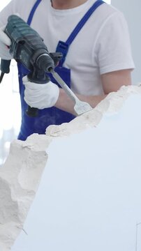 Construction worker demolishing white wall with rotary hammer drill, wearing blue overalls and protective white gloves, generating dust during renovation project, closeup view