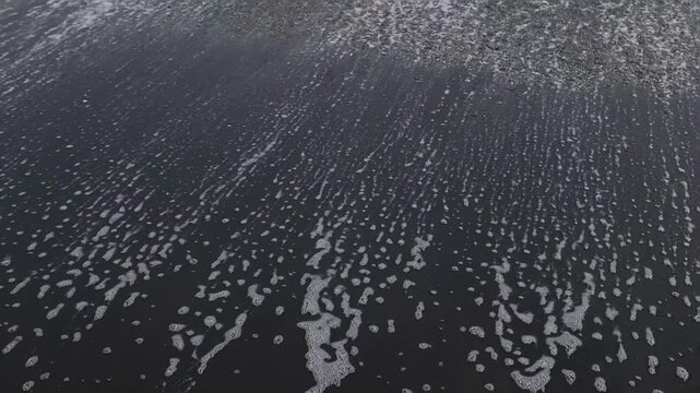 Close-up of white sea foam spreading across dark wet sand on a beach in Kamakura, Japan. Abstract coastal texture for travel, nature and ocean background themes.