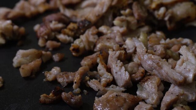 Chunks of grilled meat browning in a dark pan. The meat sizzles and browns lightly. A close-up shot reveals the texture and color. Ideal for food, recipe, or cooking videos.