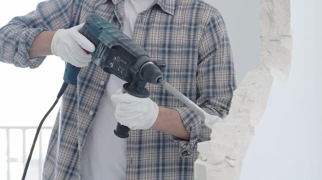 Construction worker demolishing white wall with rotary hammer drill, wearing grey checkered shirt and protective white gloves, generating dust during renovation project, closeup view