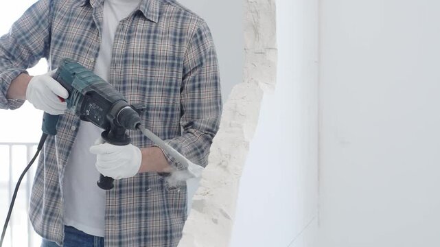 Construction worker demolishing white wall with rotary hammer drill, wearing grey checkered shirt and protective white gloves, generating dust during renovation project, closeup view
