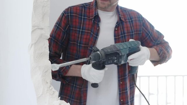 Construction worker demolishing white wall with rotary hammer drill, wearing red checkered shirt and protective white gloves, generating dust during renovation project, closeup view
