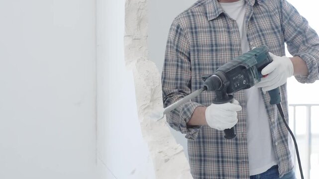 Construction worker demolishing white wall with rotary hammer drill, wearing grey checkered shirt and protective white gloves, generating dust during renovation project, closeup view